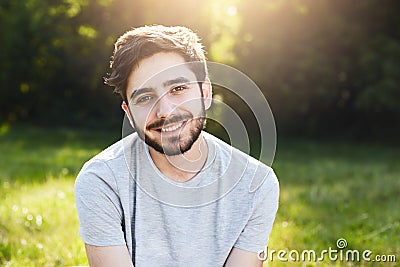 Young smiling man with dark hair, thick eyebrows, appealing eyes and beard wearing casual T-shirt sitting at greenland having good Stock Photo