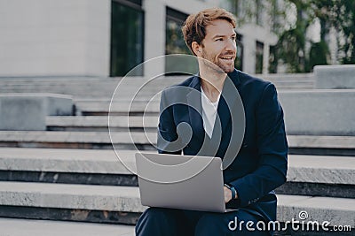 Young smiling office worker sitting outside on stairs in city center holding laptop on his lap Stock Photo