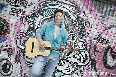 Young smiling street musician leaning on a wall with graffiti drawings and playing his guitar Editorial Stock Photo