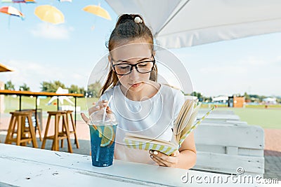 Young smiling teenager girl resting in summer outdoor cafe, girl reading book and drinking summer refreshing fruit cocktail Stock Photo