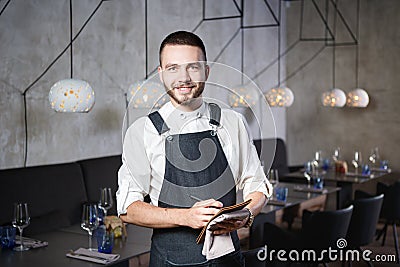 A young, smiling waiter in a restaurant, standing next to the tables with a glass of wine. Dressed in an apron, will take an order Stock Photo