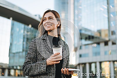 Young smiling woman in coat with coffee cup using mobile phone in evening city street Stock Photo