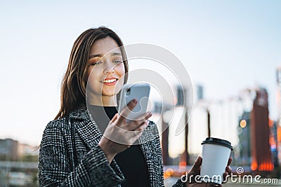 Young smiling woman in coat with coffee cup using mobile phone in evening city street Stock Photo