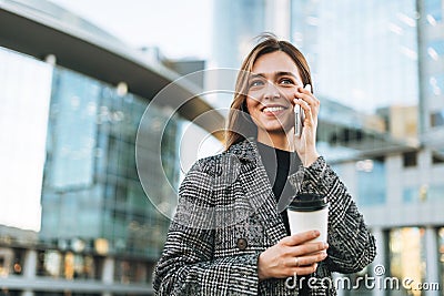 Young smiling woman in coat with coffee cup using mobile phone in evening city street Stock Photo