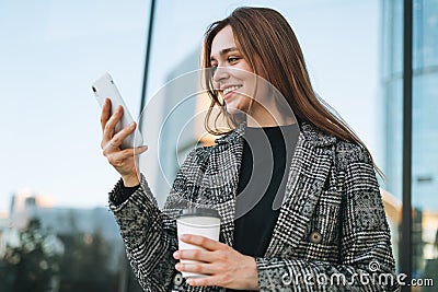 Young smiling woman in coat with coffee cup using mobile phone in evening city street Stock Photo
