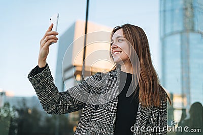 Young smiling woman in coat taling selfie on mobile phone in evening city street Stock Photo