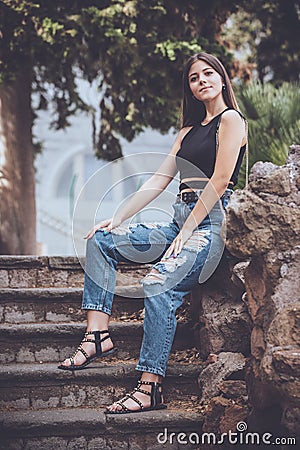 Young smiling woman jeans and accessories. Outdoors sitting on a low wall of rock on a staircase. Long hair Stock Photo