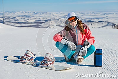 A young smiling woman sits on a mountain slope with a snowboard and a thermos Stock Photo