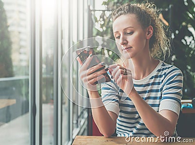 Young smiling woman in striped blouse is sitting in cafe at wooden table near window and using smartphone,reading e-book Stock Photo