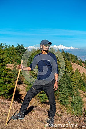 Young solo trekker with bamboo walking stick in the Himalayan hills, backdrop of glacial peaks. Uttarakhand, India. Adventurous Editorial Stock Photo