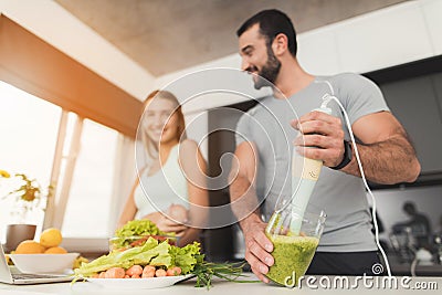 Young sports couple is preparing in the morning in the kitchen. A man is making a green vegetable cocktail. Stock Photo