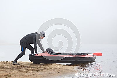 Young sportsman being ready rowing canoe, pushing boat into water, kayaker in warm clothes, padding in early fogging morning, Stock Photo