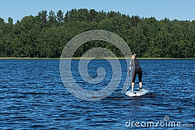 A young sporty blonde woman in the wetsuit is surfing on the sup board in the aquatory of the forest lake on bright summer day. Editorial Stock Photo