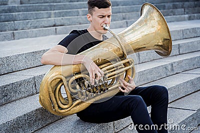 Young street musician playing tuba sitting on granite steps Stock Photo