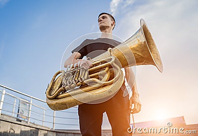 Young street musician playing tuba at the sunset sky background. Stock Photo