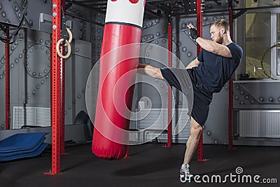 Young strong mixed martial arts fighter delivers strong side kick to punching bag. Active exercise in gym. Stock Photo