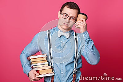 Young student in blue shirt in one tone, looks sleepy and exuasted, holds coffee near head and huge stack of books. Male tired Stock Photo
