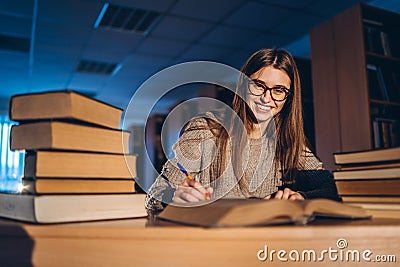 Young student in glasses preparing for the exam. Girl in the evening sits at a table in the library with a pile of books, smiling Stock Photo