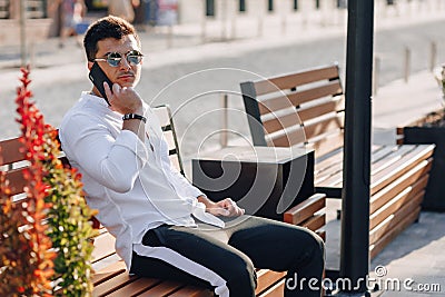 Young stylish guy in shirt with phone on bench on sunny warm day outdoors Stock Photo