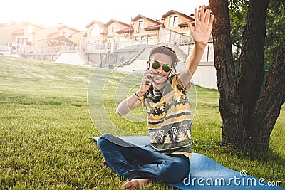 Young stylish handsome smiling man with sunglasses is sitting on grass, resting and talking on the phone. waves his hand welcoming Stock Photo