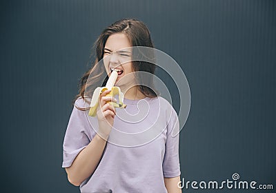 Young stylish trendy woman isolated over grey blue background. Girl biting piece of yellow ripe banana and doing weird Stock Photo
