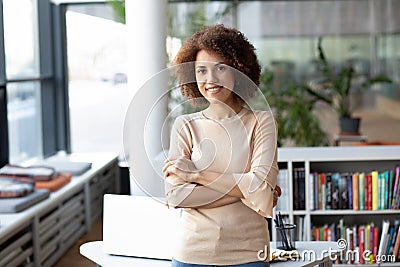 Young successful African American woman entrepreneur stands with crossed arms near a desk in a modern office, looking at the Stock Photo