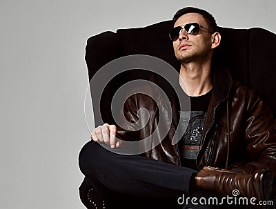 Young successful rich man in black t-shirt brown leather jacket and brown leather boots sits in big armchair Stock Photo