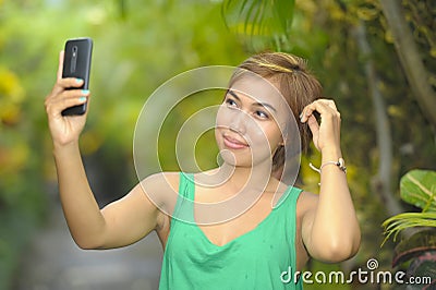 Young sweet and pretty Asian woman holding mobile phone taking selfie picture in tropical jungle smiling happy posing cool in soci Stock Photo