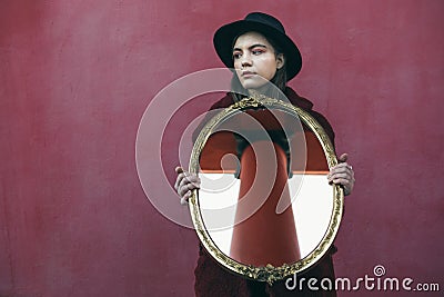 Young teen girl in hat holding mirror in front of red wall. the mirror reflects the column of the building opposite Stock Photo