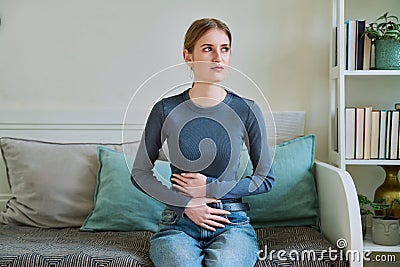 Young teenage female experiencing abdominal pain, sitting on couch at home Stock Photo