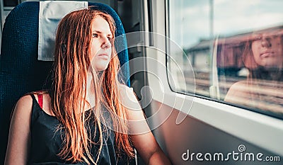 Young teenager redhead girl with long hair looks out the window while sitting in the train. Stock Photo