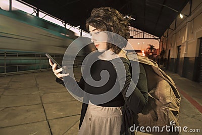 Young teenager woman with backpack alone at train station cheking smart phone at evening light Stock Photo