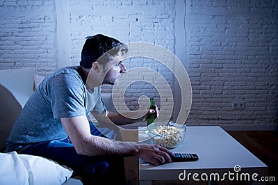 Young television addict man sitting on home sofa watching TV eating popcorn and drinking beer bottle Stock Photo