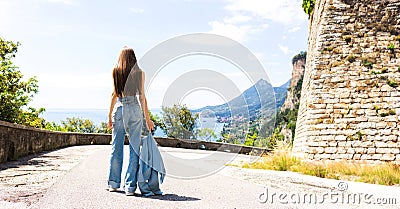 A young and thin girl walks on a beautiful road facing the lake and the mountain Stock Photo