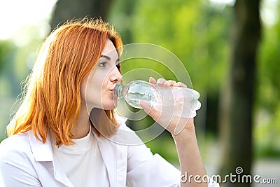 Young thirsty redhead woman drinking water from a bottle in summer park Stock Photo
