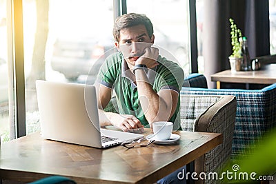 Young thoughtful quizzical businessman in green t-shirt sitting with laptop, hand on chin looking away and thinking what to do Stock Photo