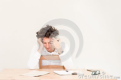 Young, tired Caucasian freelancer man sitting in his home office at the table in front of empty clear wall. Stock Photo