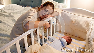 Young tired and exhausted mother fallen asleep while rocking crib of her newborn baby at night. Concept of sleepless Stock Photo