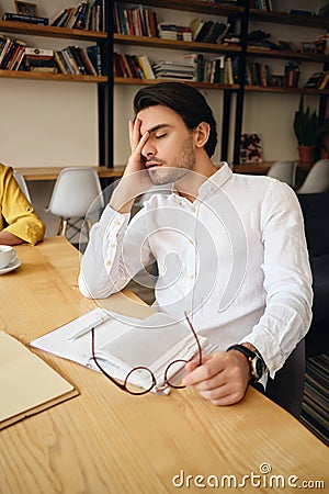 Young tired man sitting at the table with notepad leaning head on hand sleeping at work in modern office Stock Photo