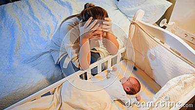 Young tired mother suffering from depression lookin on her sleepless newborn baby in crib at night. Maternal depression Stock Photo