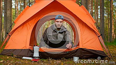 Young traveler guy talks to client via video link via laptop while sitting in nature in tent. Man talks to his boss on Stock Photo
