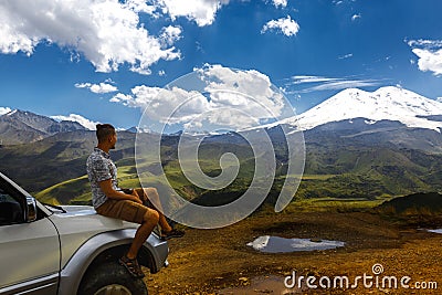 Young Traveler Man Sits On Car And Enjoys View Of Mountains In Summer. Elbrus Region, North Caucasus, Russia Stock Photo