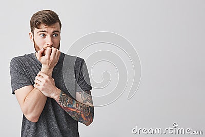 Young unhappy bearded tattooed european man with fashionable short haircut in casual gray shirt gnawing fingers, looking Stock Photo