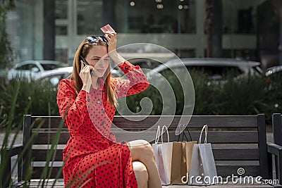Young upset Caucasian woman in red dress sitting on a bench talking on the phone to clear up a charge on her credit card Stock Photo