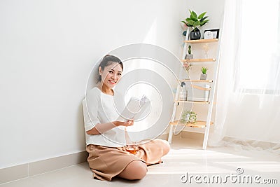 Young Vietnamese girl drinkng tea and reading a book while sitting on the floor Stock Photo