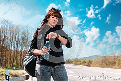 A young waiting caucasian woman looks at her wristwatch. Countryside road on the background. The concept of waiting Stock Photo