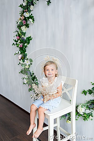 A young warm smiling girl is sitting on the wooden white chair near the wall with flowers wreath. Stock Photo