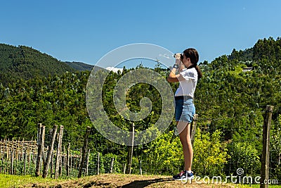 Young white Caucasian girl standing in shorts looking through a pair of binoculars at the mountainside next to an orchard on a Stock Photo