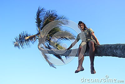 A young white man climbed onto a palm tree and sits on a trunk amid a bright blue sky and palm Stock Photo