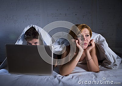 young wife upset unsatisfied and frustrated in bed while husband work on computer laptop ignoring her Stock Photo